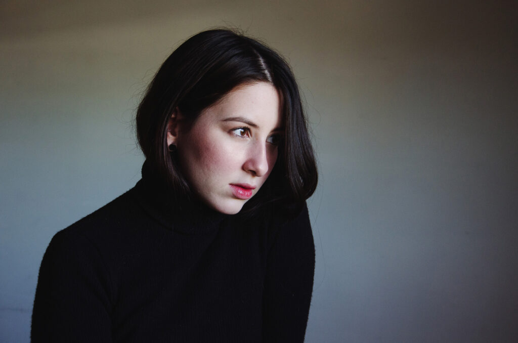 Gabriela Inzunza gazes off to the side against a muted background. She wears a black turtleneck and her dark hair cascades down one side of her face.